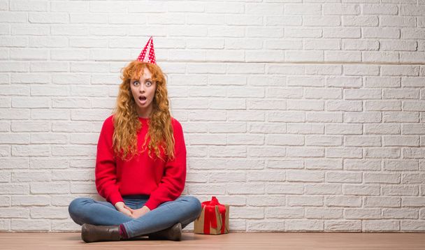 Jeune femme rousse assise sur un mur de briques portant un chapeau d'anniversaire effrayé en état de choc avec un visage surprise, effrayé et excité par l'expression de la peur
 - Photo, image