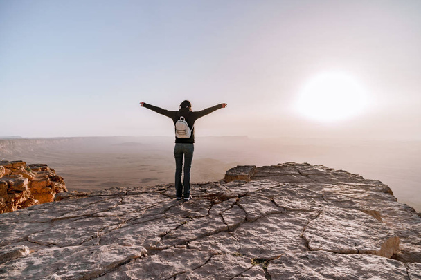 Одна молодая девушка с рюкзаком в пустыне Исраэль Негев любуется видом восхода солнца. Молодая женщина стоит на краю утёса парка Махтеш Рамон
. - Фото, изображение