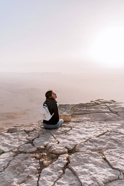 Alone young girl with backpack in israel negev desert admires the view of sunrise. Young female person stands on the edge of the cliff of makhtesh ramon park. - Photo, Image