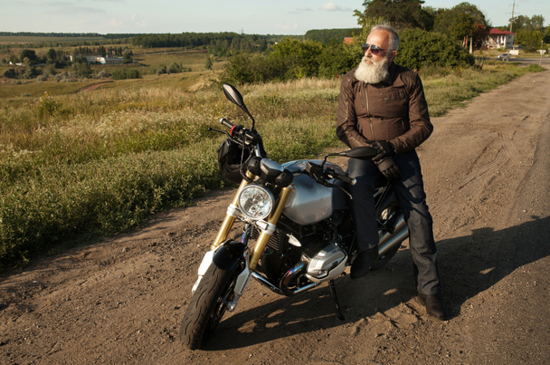 Homem velho motociclista vestindo uma jaqueta de couro e óculos de sol sentados em sua moto
. - Foto, Imagem