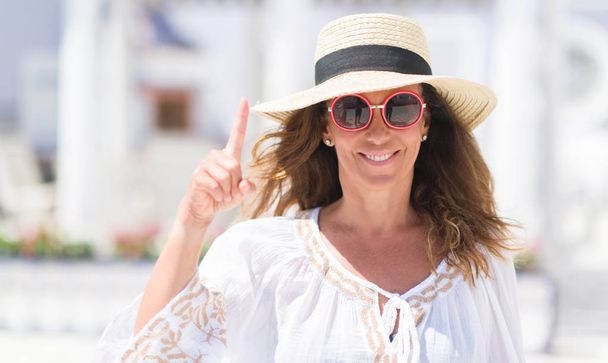 Donna bruna di mezza età con occhiali da sole e cappello estivo sorpresa da un'idea o domanda che punta il dito con il viso felice, numero uno
 - Foto, immagini