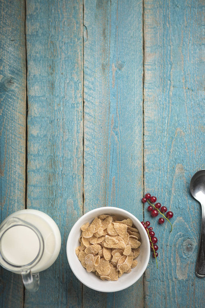 Organic corn or oat or cereal or barley flakes in blue porcelain bowl with red currant and jug of milk on old wooden table - Fotoğraf, Görsel