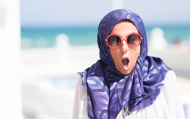 Mujer árabe morena de mediana edad junto a la piscina con gafas de sol asustada en shock con una cara sorpresa, asustada y excitada con expresión de miedo
 - Foto, Imagen