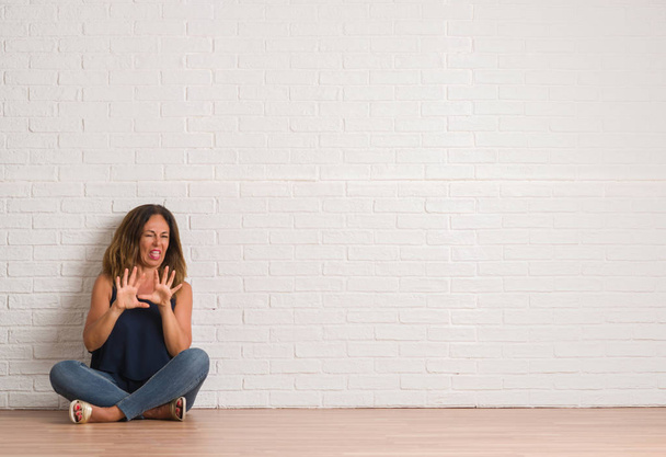 Hispanische Frau mittleren Alters, die auf dem Boden über der weißen Backsteinwand sitzt, ist angewidert, ungehalten und ängstlich, weil sie Ekelgesicht tut, weil sie eine Abneigung dagegen hat. mit erhobenen Händen. Ärgerliches Konzept. - Foto, Bild