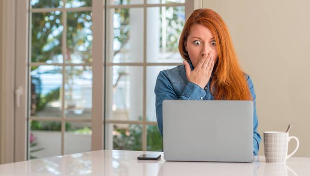 Redhead woman using computer laptop at home cover mouth with hand shocked with shame for mistake, expression of fear, scared in silence, secret concept - Фото, изображение