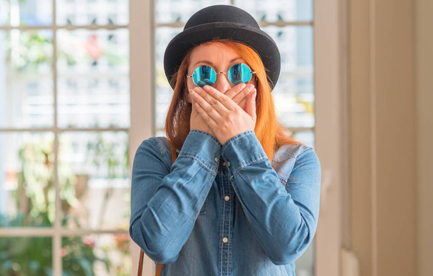 Stylish redhead woman wearing bowler hat and sunglasses shocked covering mouth with hands for mistake. Secret concept. - Photo, Image