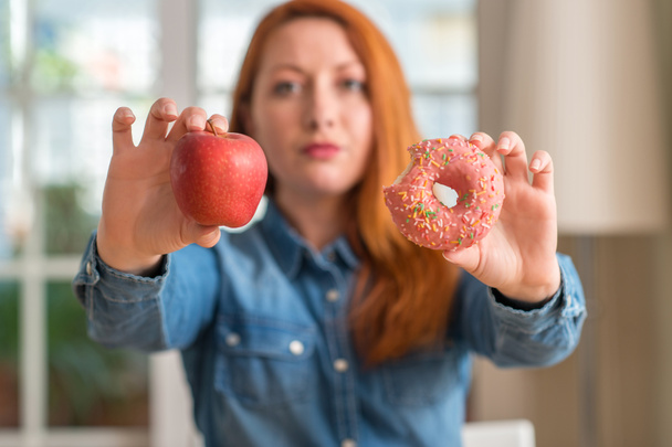 Pelirroja mujer elige entre manzana y donut con una expresión de confianza en la cara inteligente pensando en serio
 - Foto, imagen