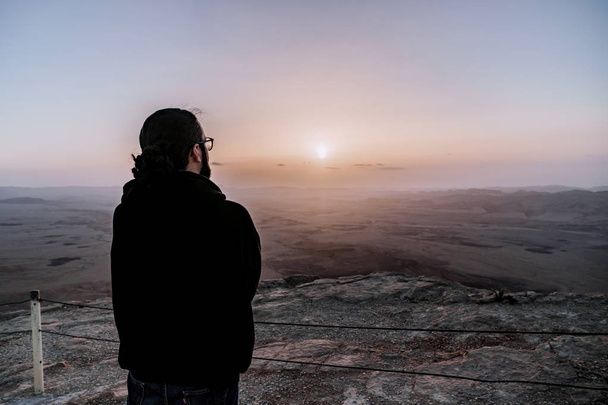 Один человек в пустыне Исраэль Негев восхищается видом восхода солнца. Молодой мужчина стоит на краю утёса
 - Фото, изображение