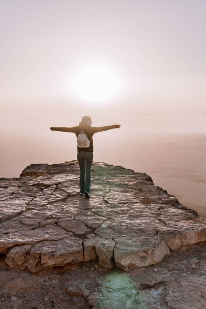 Одна молода дівчина з рюкзаком в пустелі Ізраїль Негев захоплюється видом на схід сонця. Молода жінка стоїть на краю скелі парку барана Махтеш
. - Фото, зображення