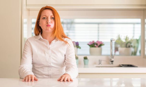 Redhead woman at kitchen puffing cheeks with funny face. Mouth inflated with air, crazy expression. - 写真・画像