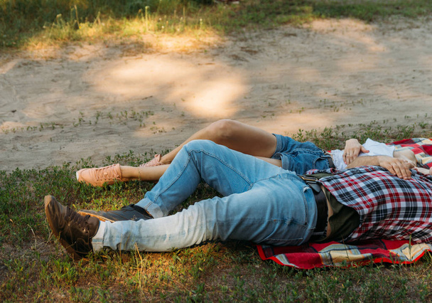 loving couple lie, lie down on a red plaid in the woods and embrace.the couple is kissing.on the man dressed in a plaid shirt and jeans. a girl hugs a man, dressed in a white sweater and denim shorts. summer rest - Foto, afbeelding