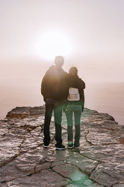 Alone young couple of man and women with backpack in israel negev desert admires the view of sunrise. Young pair stands on the edge of the cliff of makhtesh ramon park. - Foto, Imagem