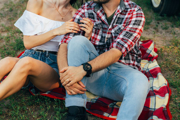 loving couple sits on a red plaid in the woods and embrace.the couple is kissing.on the man's arm is a black watch, dressed in a plaid shirt and jeans. a girl hugs a man, dressed in a white sweater and denim shorts. summer rest - Fotoğraf, Görsel