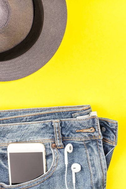 Flat lay of blue jeans, grey hat and smartphone headphones on bold yellow paper background with copy space. Overhead view of woman casual outfit. Trendy hipster look top view - Foto, imagen