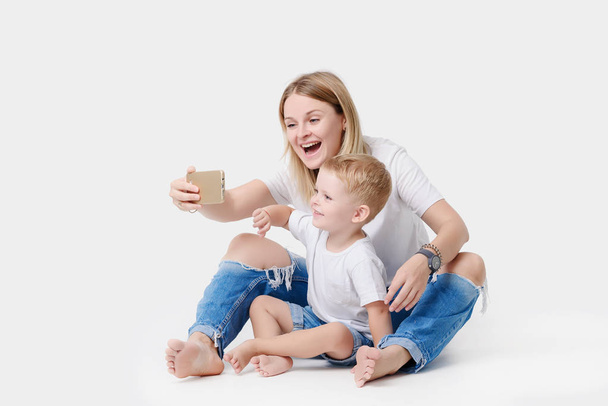 Happy young mother playing with her young son on a white isolated background. Take pictures and videos on your mobile phone, make selfie - Φωτογραφία, εικόνα