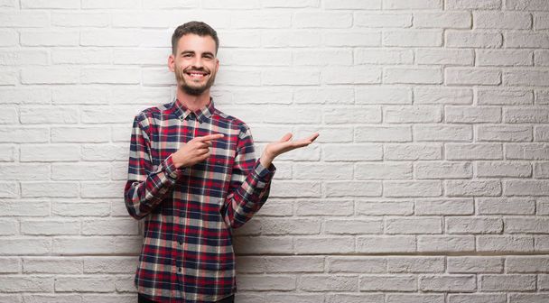 junger erwachsener Mann über weiße Backsteinmauer sehr glücklich mit Hand und Finger zeigend - Foto, Bild
