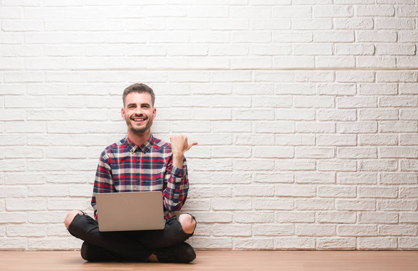 Mladý dospělý muž nad cihlovou zeď přenosném počítači najeďte a ukazuje s palcem na stranu s úsměvem a šťastný obličej - Fotografie, Obrázek