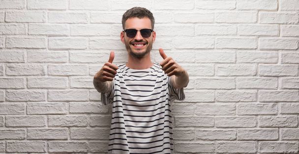 Giovane uomo adulto con gli occhiali da sole in piedi sopra muro di mattoni bianchi che approva facendo gesto positivo con la mano, pollici in su sorridente e felice per il successo. Guardando la macchina fotografica, il gesto del vincitore
. - Foto, immagini