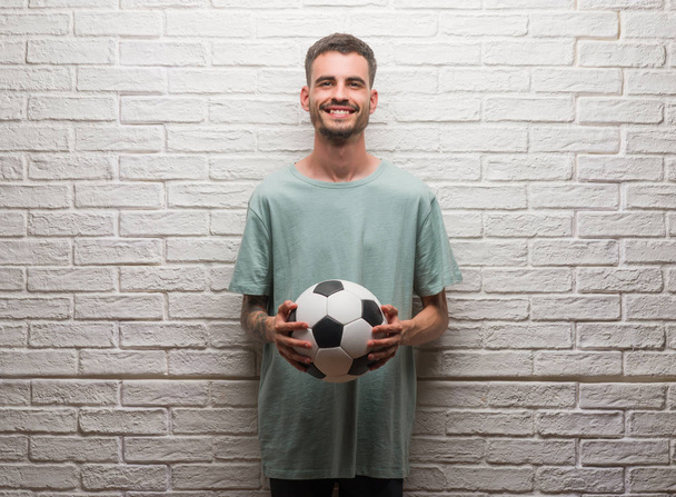Giovane uomo adulto che tiene il pallone da calcio in piedi sopra il muro di mattoni bianchi con una faccia felice in piedi e sorridente con un sorriso fiducioso che mostra i denti
 - Foto, immagini