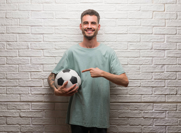 Jovem adulto homem segurando bola de futebol em pé sobre parede de tijolo branco muito feliz apontando com a mão e o dedo
 - Foto, Imagem
