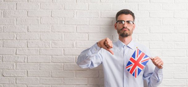 Hombre joven adulto sobre pared de ladrillo sosteniendo la bandera del Reino Unido con la cara enojada, signo negativo que muestra aversión con los pulgares hacia abajo, concepto de rechazo
 - Foto, Imagen