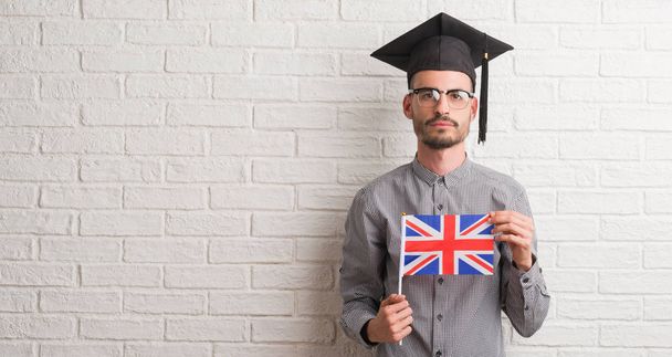 Hombre adulto joven sobre la pared de ladrillo con gorra de graduación sosteniendo bandera del Reino Unido con una expresión de confianza en la cara inteligente pensando en serio
 - Foto, imagen