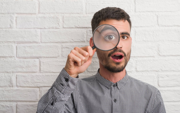 Young adult man over brick wall using magnifying glass scared in shock with a surprise face, afraid and excited with fear expression - Photo, Image