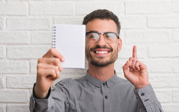 Young adult man holding notebook standing over white brick wall surprised with an idea or question pointing finger with happy face, number one - Fotoğraf, Görsel