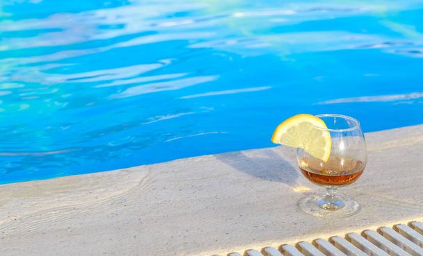 Blue pool water, a glass of flavored cognac with a slice of lemon at the water's edge - Photo, Image