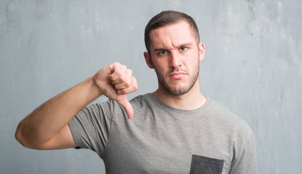 Homem caucasiano jovem sobre parede grunge cinza com rosto irritado, sinal negativo mostrando desgosto com os polegares para baixo, conceito de rejeição
 - Foto, Imagem