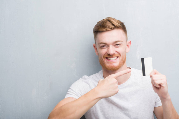 junger rothaariger Mann über graue Grunge-Wand mit Kreditkarte sehr glücklich zeigt mit Hand und Finger - Foto, Bild