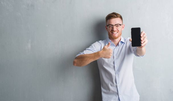 Jonge roodharige man over grijs grunge muur tonen leeg scherm van een smartphone blij met de grote glimlach doen ok teken, duim omhoog met vingers, uitstekende teken - Foto, afbeelding