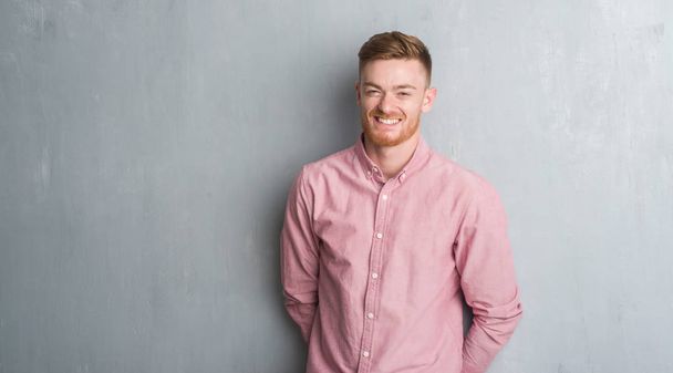 Young redhead man over grey grunge wall wearing pink shirt with a happy and cool smile on face. Lucky person. - Photo, Image