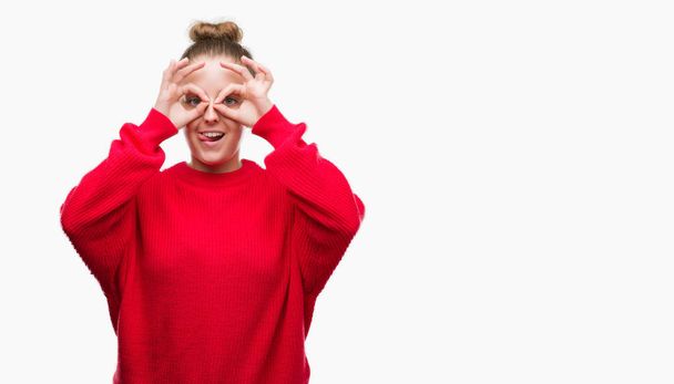 Young blonde woman wearing bun and red sweater doing ok gesture like binoculars sticking tongue out, eyes looking through fingers. Crazy expression. - Foto, immagini