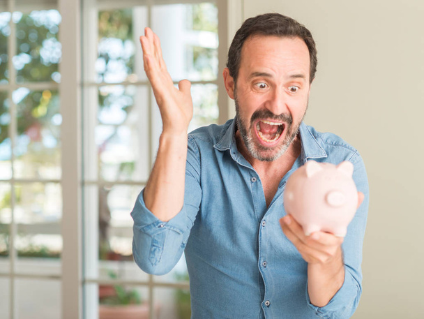 Middle age man save money on piggy bank very happy and excited, winner expression celebrating victory screaming with big smile and raised hands - Foto, Bild