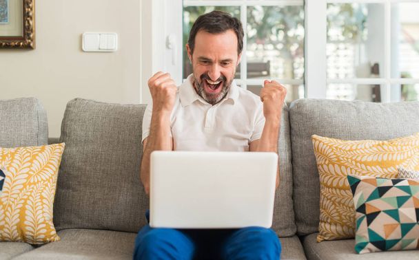Мужчина средних лет, используя ноутбук на диване кричать гордо и праздновать победу и успех очень взволнован, аплодируя эмоции
 - Фото, изображение