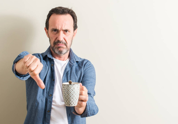 Hombre mayor bebiendo café en una taza con la cara enojada, signo negativo que muestra aversión con los pulgares hacia abajo, concepto de rechazo
 - Foto, Imagen