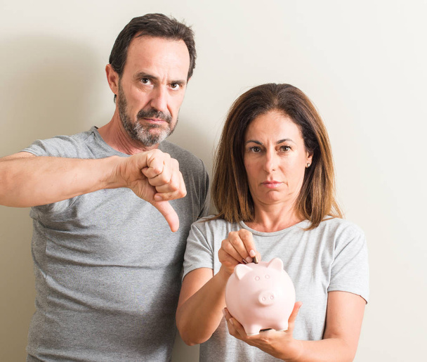 Para w średnim wieku, Kobieta i mężczyzna, trzymając Skarbonka z gniewną twarz, ujemna znak wyświetlone niechęć z kciuk w dół, odrzucenie koncepcji - Zdjęcie, obraz