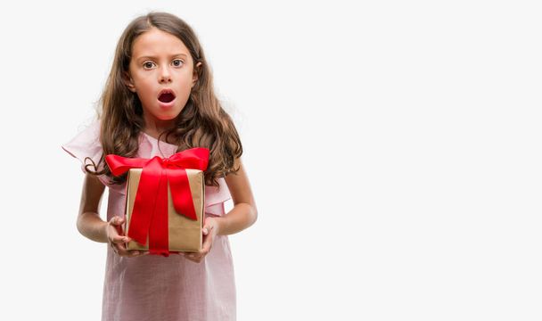 brünettes hispanisches Mädchen, das ein Geschenk in der Hand hält, erschrocken mit einem Überraschungsgesicht, ängstlich und aufgeregt mit Angstausdruck - Foto, Bild