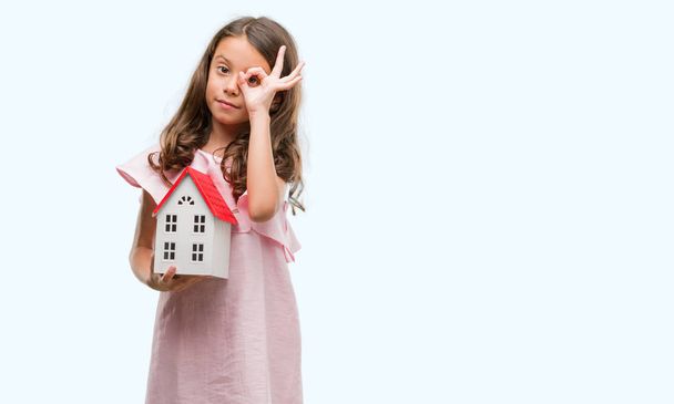 Brunette hispanic girl holding little house with happy face smiling doing ok sign with hand on eye looking through fingers - Foto, immagini