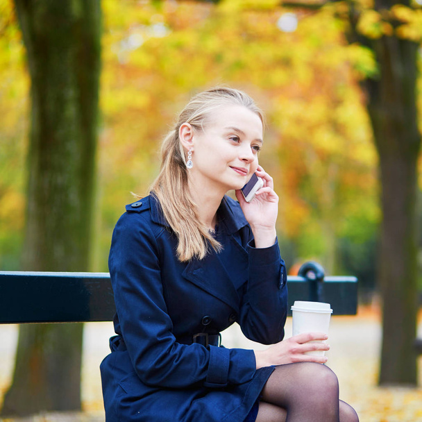 Beautiful young woman drinking coffee and speaking on the phone in the Luxembourg garden of Paris on a fall day - Photo, Image