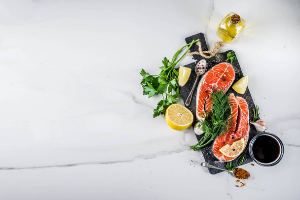 Raaka lohi kala pihvejä sitruuna, yrttejä, oliiviöljy, valmis grilli, liuskekivi leikkuulauta, valkoinen marmori tausta kopio tilaa edellä
 - Valokuva, kuva