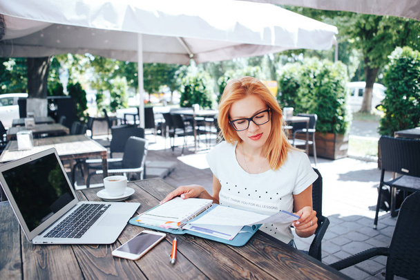 Młoda Rudowłosa kobieta patrząc w notesie w letniej kawiarni. Młoda kobieta siedzi w kawiarni i działa - Zdjęcie, obraz