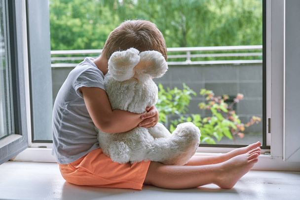 boy of two years sitting by the window and hugs a toy Bunny. rainy weather, waiting for dad to come home from work - 写真・画像