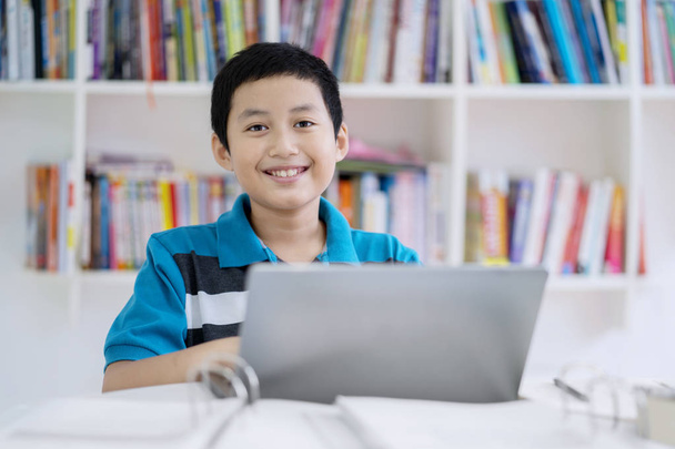 Picture of Asian preteen student looks happy while using a laptop in the library - Фото, изображение