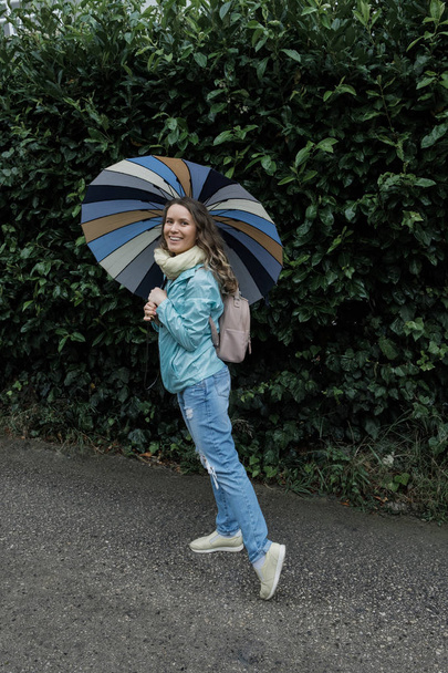 Νεαρός χαμογελαστό ξανθιά γυναίκα περπάτημα με ομπρέλα στη βροχή το φθινόπωρο ή την άνοιξη στο πάρκο - Φωτογραφία, εικόνα