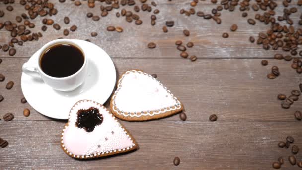 Food art. Good morning concept. cup of coffee and 2 heart-shaped gingersnaps are on wooden background. Coffee beans fall down in slow motion - Footage, Video