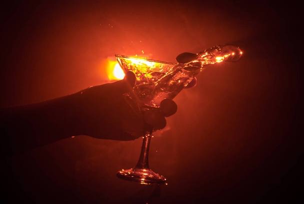 Martini cocktail glass in hand splashing on dark toned smoky background or colorful cocktail in glass with splashes and olives. Party club entertainment. Mixed light. Selective focus - Photo, image