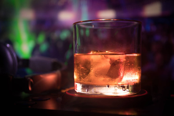 Glass with whisky with ice cube inside on dj controller at nightclub. Dj Console with club drink at music party in nightclub with disco lights. Selective focus - Фото, изображение