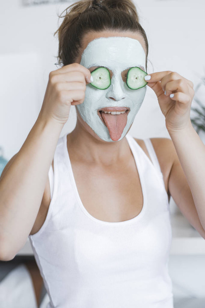 Young Pretty Blonde woman with facial mask posing with cucumber at home. Wellbeing concept, Morning skin care routine. - Foto, imagen
